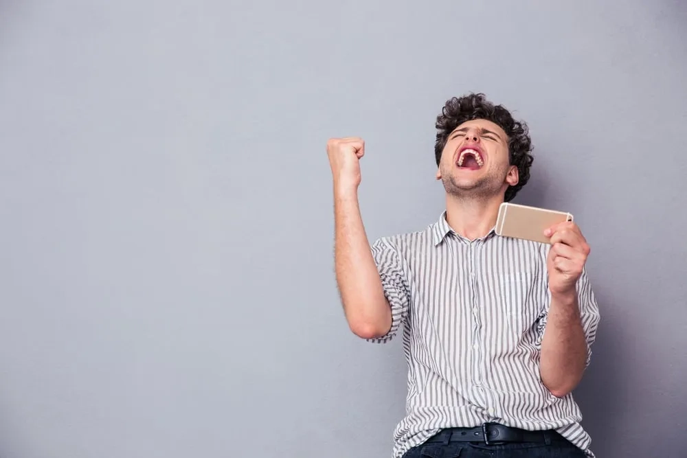 Happy man holding smartphone and celebrating his success over gray background
